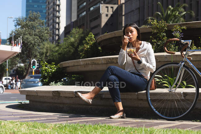 Mujer afroamericana tomando un aperitivo mientras está sentada en el parque corporativo. estilo de vida concepto de vida durante el coronavirus covid 19 pandemia. - foto de stock