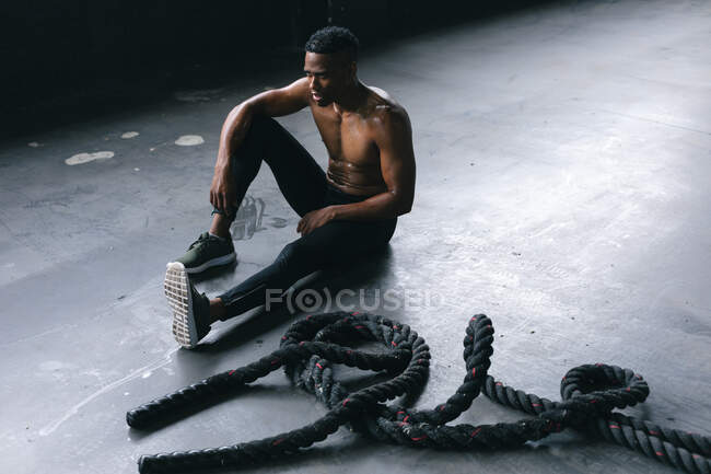 Homme afro-américain portant des vêtements de sport assis au repos après avoir lutté contre des cordes dans un bâtiment urbain vide. forme physique urbaine mode de vie sain. — Photo de stock