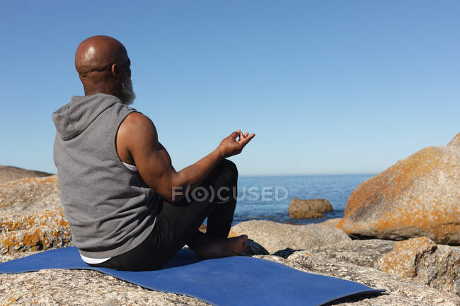 Fit senior african american man sitting on rocky coast meditating. healthy retirement outdoor fitness lifestyle. — Stock Photo