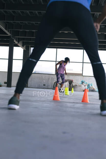 Femme afro-américaine faisant du slalom avec un ballon de football dans un bâtiment urbain vide. homme l'acclamant. forme physique urbaine mode de vie sain. — Photo de stock