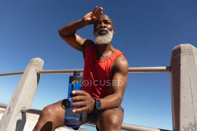 Se adapta al hombre afroamericano mayor haciendo ejercicio sentado al sol sosteniendo una botella de agua. retiro saludable estilo de vida fitness al aire libre. - foto de stock