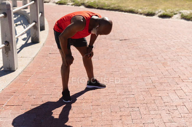 Ajustez l'homme afro-américain senior portant des écouteurs s'exerçant au soleil reprenant son souffle. retraite saine sport forme physique extérieure mode de vie. — Photo de stock