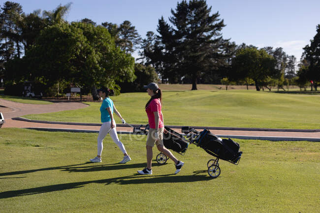 Duas mulheres caucasianas a atravessar o campo de golfe a puxar sacos de golfe sobre rodas. esporte lazer hobbies golfe saudável ao ar livre estilo de vida. — Fotografia de Stock