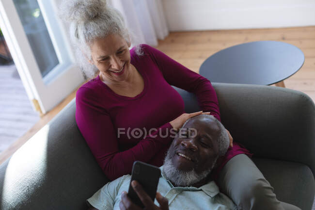 Sénior casal misto abraçando olhando para o smartphone juntos na sala de estar. ficar em casa em auto-isolamento durante o confinamento de quarentena. — Fotografia de Stock