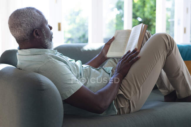 Старший африканский американец лежит на диване и читает книгу. оставаться дома в изоляции во время карантинной изоляции. — стоковое фото