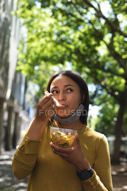Mujer afroamericana con mascarilla baja comiendo algo en la calle. estilo de vida concepto de vida durante el coronavirus covid 19 pandemia. - foto de stock