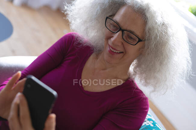Eine ältere Frau sitzt mit ihrem Smartphone auf der Couch im Wohnzimmer. Während der Quarantäne zu Hause bleiben und sich selbst isolieren. — Stockfoto