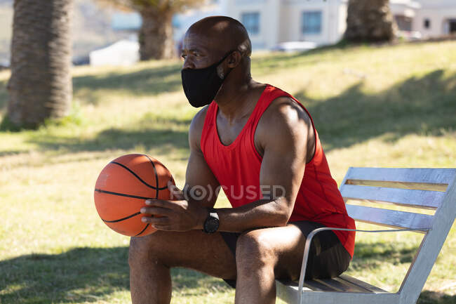 Fit Senior afrikanisch-amerikanischer Mann mit Gesichtsmaske sitzt im Park und hält Basketball. gesunder Ruhestand Outdoor-Fitness-Lebensstil Hygiene während Coronavirus covid 19 Pandemie. — Stockfoto