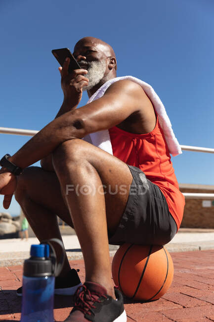 Ajuste hombre afroamericano senior sentado en el baloncesto en el sol hablando en el teléfono inteligente. saludable retiro tecnología comunicación al aire libre fitness estilo de vida. - foto de stock