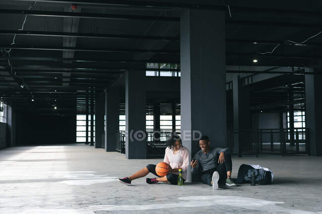 Африканские американец мужчина и женщина сидят в пустом городском здании и отдыхают после игры в баскетбол. используя смартфон и смеясь. здоровый образ жизни. — стоковое фото