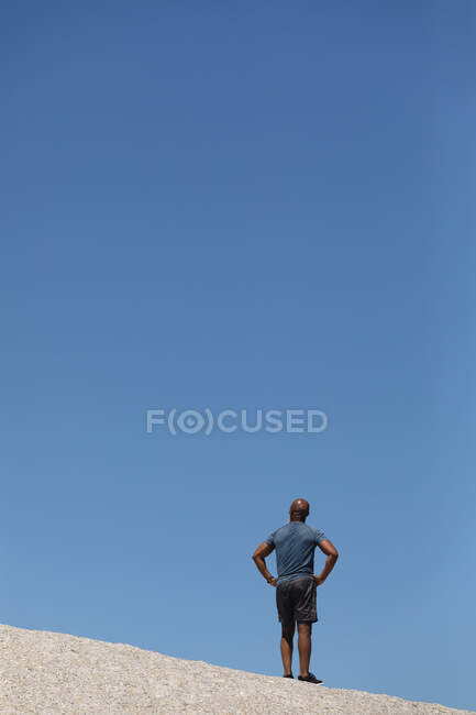 Vista trasera del hombre afroamericano mayor en forma de pie sobre roca contra el cielo azul. retiro saludable estilo de vida fitness al aire libre. - foto de stock