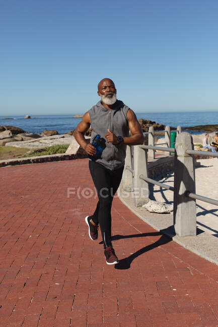 Ajuste hombre afroamericano senior ejerciendo correr en la ruta costera. retiro saludable estilo de vida fitness al aire libre. - foto de stock