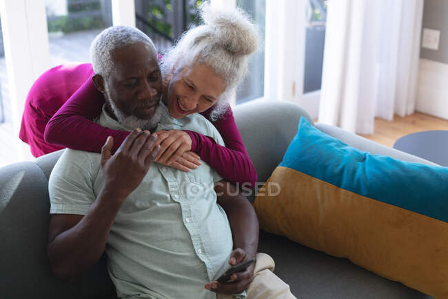 Sénior casal misto abraçando olhando para o smartphone juntos na sala de estar. ficar em casa em auto-isolamento durante o confinamento de quarentena. — Fotografia de Stock