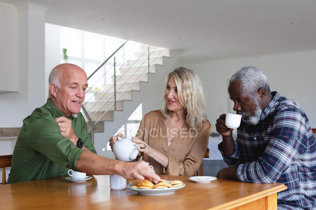 Senior caucasian and african american people sitting by table drinking tea at home. senior retirement lifestyle friends socializing. — Stock Photo