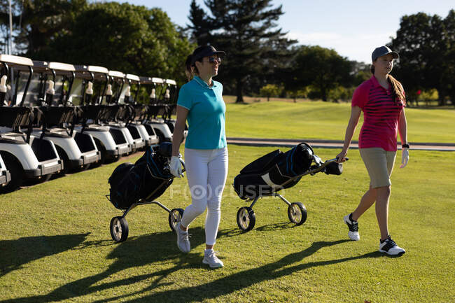 Due donne caucasiche che attraversano il campo da golf tirando sacchi da golf su ruote. sport tempo libero hobby golf sano stile di vita all'aperto. — Foto stock