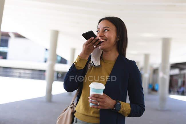 Mujer afroamericana con taza de café hablando en smartphone en la calle. estilo de vida durante la pandemia de coronavirus covid 19. - foto de stock