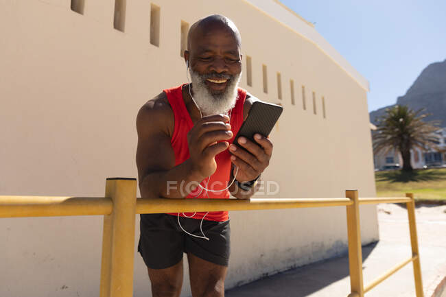 Ajuste hombre afroamericano senior usando auriculares usando teléfono inteligente en la calle soleada sonriendo. saludable retiro tecnología comunicación al aire libre fitness estilo de vida. - foto de stock