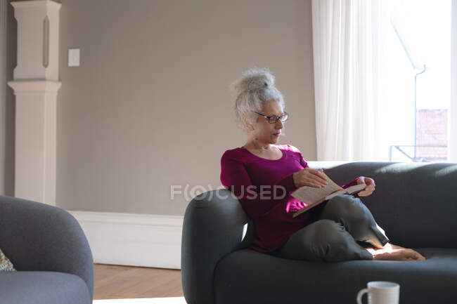 Старшая белая женщина, сидящая на диване в гостиной и читающая книгу. оставаться дома в изоляции во время карантинной изоляции. — стоковое фото