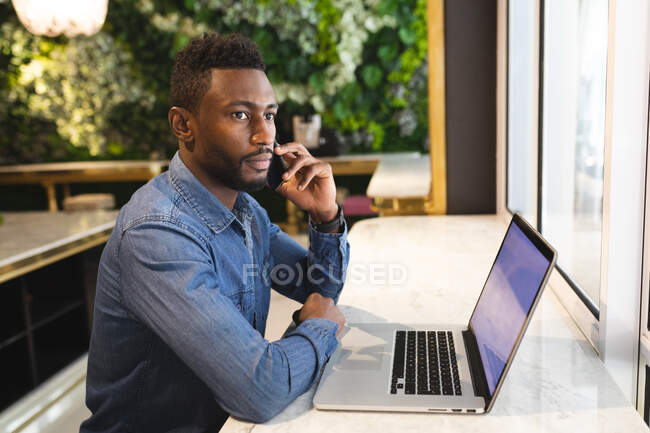 Homem afro-americano sentado em um café falando usando um smartphone e usando um laptop. homem de negócios em movimento na cidade. — Fotografia de Stock