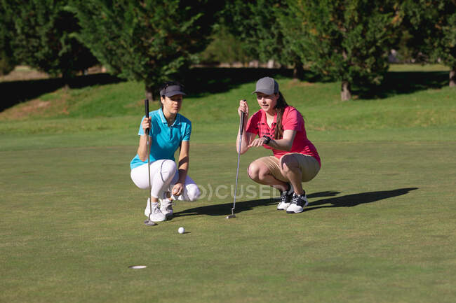 Dos mujeres caucásicas jugando al golf en cuclillas cerca del hoyo hablando. deporte ocio aficiones golf estilo de vida al aire libre saludable. - foto de stock
