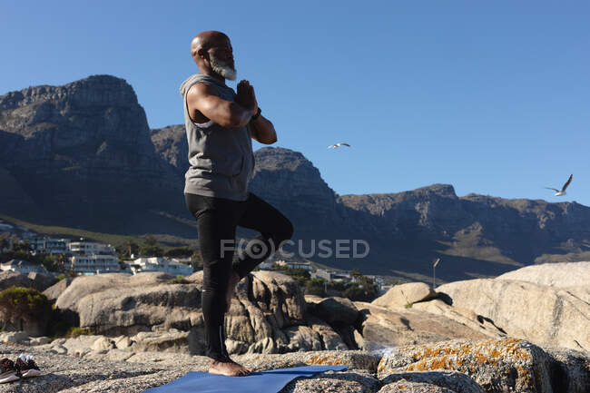 Fit senior african american man standing on rocky coast doing yoga. healthy retirement outdoor fitness lifestyle. — Stock Photo