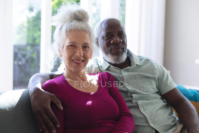 Senior coppia di razza mista abbracciando guardando la fotocamera e sorridendo in soggiorno. stare a casa in isolamento durante l'isolamento in quarantena. — Foto stock