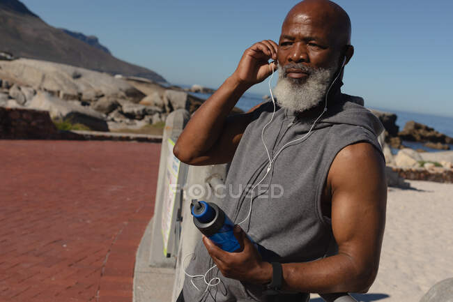 Ajuste o homem americano africano sênior pelo mar usando fones de ouvido que prendem a garrafa de água. tecnologia de aposentadoria saudável comunicação ao ar livre fitness lifestyle. — Fotografia de Stock