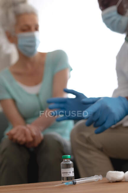 Médico do sexo masculino afro-americano preparando a vacinação covid 19 para a mulher caucasiana sênior em casa. cuidados de saúde e higiene em casa em auto-isolamento durante o bloqueio de quarentena. — Fotografia de Stock