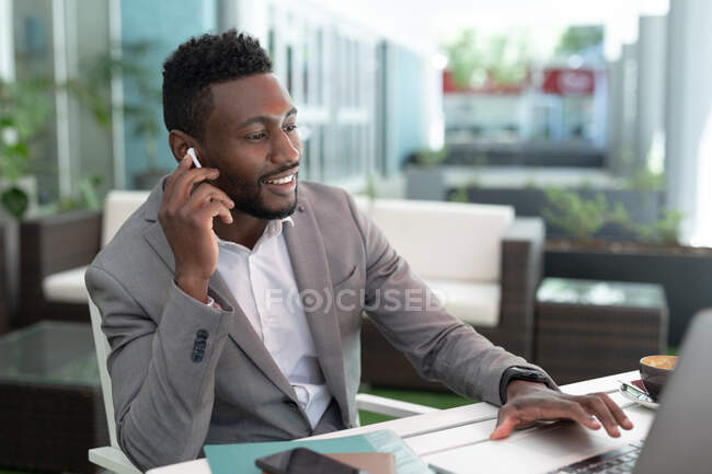Hombre de negocios afroamericano sentado en un café usando un portátil y hablando en un teléfono inteligente. hombre de negocios en la salida en la ciudad. - foto de stock