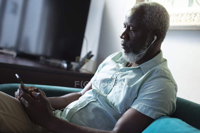 Homme afro-américain âgé assis dans le salon en utilisant un smartphone écoutant de la musique sur des écouteurs. rester à la maison en isolement personnel pendant le confinement en quarantaine. — Photo de stock