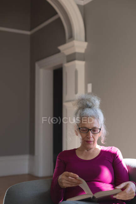 Mulher caucasiana sênior sentada no sofá na sala de estar lendo livro. ficar em casa em auto-isolamento durante o confinamento de quarentena. — Fotografia de Stock