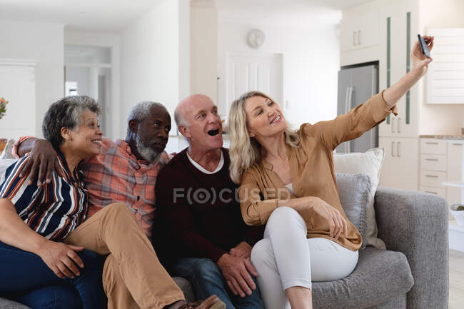Старшие кавказские и африканские американские пары сидят на диване и делают селфи дома. Друзья на пенсии общаются. — стоковое фото