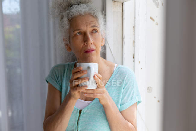 Eine ältere kaukasische Frau steht am Fenster und trinkt zu Hause Kaffee. Während der Quarantäne zu Hause bleiben und sich selbst isolieren. — Stockfoto