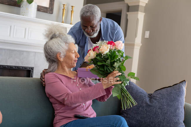 Senior mixte couple dans le salon homme donnant des fleurs femme. rester à la maison en isolement personnel pendant le confinement en quarantaine. — Photo de stock