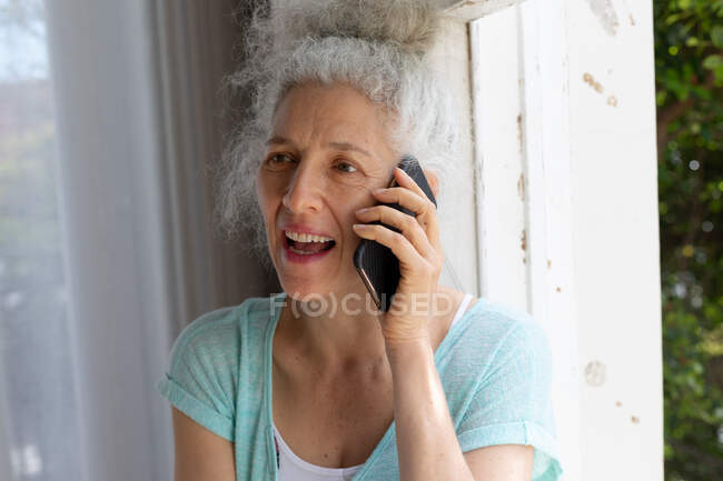 Mujer caucásica mayor de pie junto a la ventana hablando en el teléfono inteligente en casa. permanecer en casa en aislamiento durante el bloqueo de cuarentena. - foto de stock