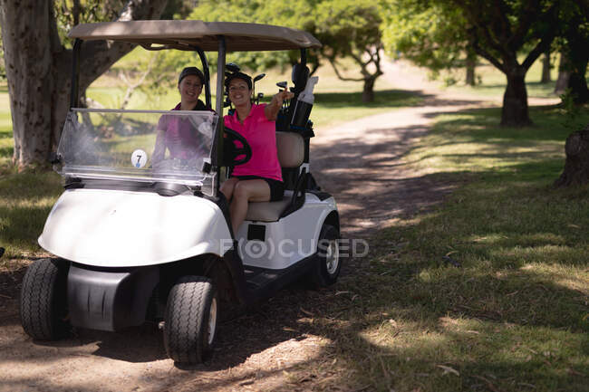 Dos mujeres caucásicas conduciendo un carrito de golf con palos en la espalda en el campo de golf. concepto de deporte y estilo de vida activo. - foto de stock