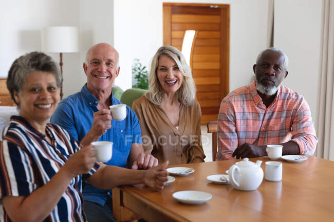 Parejas mayores caucásicas y afroamericanas sentadas junto a la mesa tomando té en casa. todos mirando a la cámara y sonriendo. senior retiro estilo de vida amigos socializar. - foto de stock