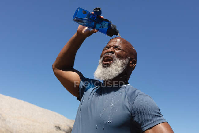 Ajustez l'homme afro-américain senior exerçant le refroidissement avec la bouteille d'eau contre le ciel bleu. retraite saine mode de vie en plein air fitness. — Photo de stock