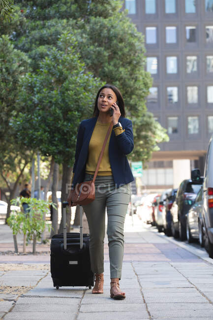Afroamerikanerin mit Trolleytasche, die auf der Straße mit dem Smartphone spricht. Lebensstil während Coronavirus-Covid 19 Pandemie. — Stockfoto