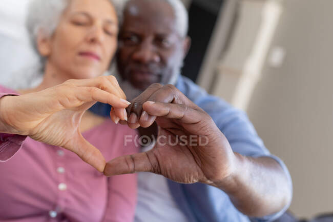 Senior coppia di razza mista in salotto abbracciando facendo una forma di cuore con le mani. stare a casa in isolamento durante l'isolamento in quarantena. — Foto stock