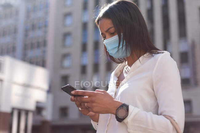 Mujer afroamericana con máscara facial usando smartphone en la calle. estilo de vida concepto de vida durante el coronavirus covid 19 pandemia. - foto de stock