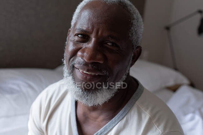 Porträt eines älteren afrikanisch-amerikanischen Mannes, der auf dem Bett sitzt, in die Kamera schaut und lächelt. Während der Quarantäne zu Hause bleiben und sich selbst isolieren. — Stockfoto