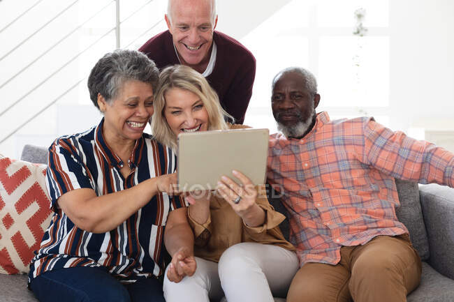 Старшие кавказские и африканские американские пары сидят на диване, используя цифровые планшеты дома. Друзья на пенсии общаются. — стоковое фото