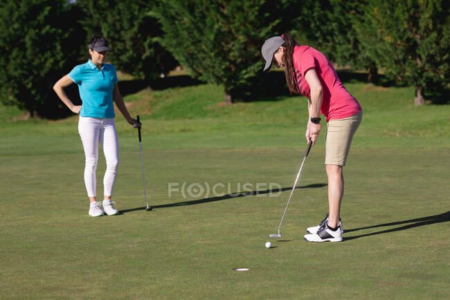 Dos mujeres caucásicas jugando al golf una disparando al hoyo. deporte ocio aficiones golf estilo de vida al aire libre saludable. - foto de stock