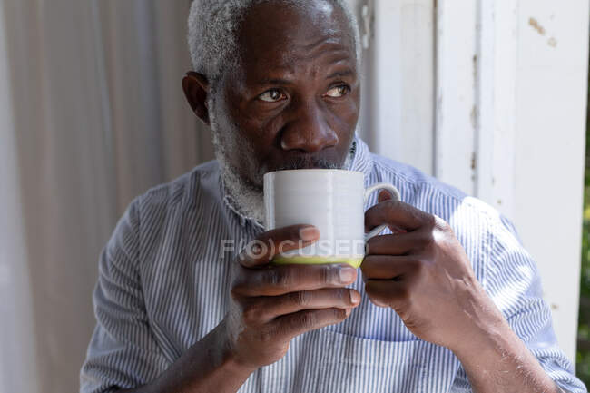 Uomo anziano afroamericano che sta vicino alla finestra a bere caffe 'a casa. stare a casa in isolamento durante l'isolamento in quarantena. — Foto stock