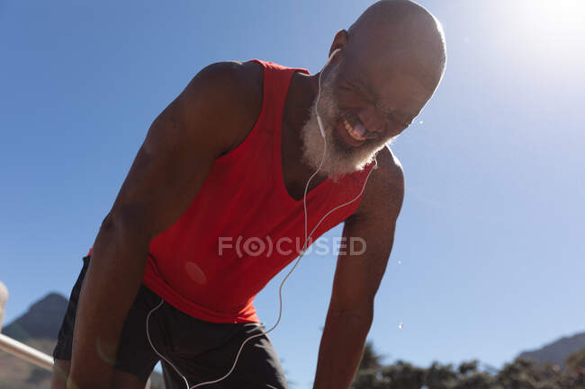 Se encaixa americano africano sênior usando fones de ouvido sorrindo contra o céu azul ensolarado. tecnologia de aposentadoria saudável comunicação ao ar livre fitness lifestyle. — Fotografia de Stock