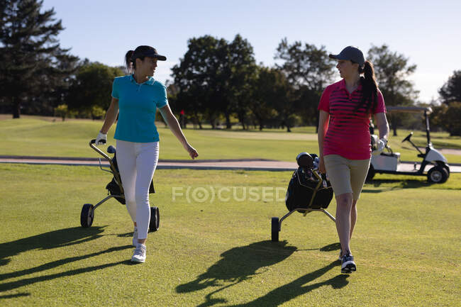 Zwei kaukasische Frauen laufen über den Golfplatz und reden und ziehen Golftaschen auf Rädern. Sport Freizeit Hobbys Golf gesunder Lebensstil im Freien. — Stockfoto