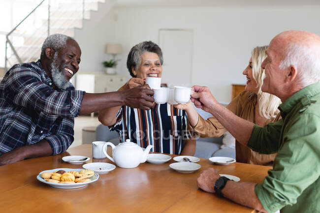 Parejas mayores caucásicas y afroamericanas sentadas junto a la mesa tomando té en casa. senior retiro estilo de vida amigos socializar. - foto de stock