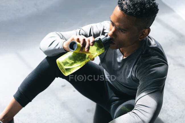 Afroamerikanischer Mann sitzt in einem leeren städtischen Gebäude und ruht sich aus, nachdem er Basketball gespielt und Wasser getrunken hat. urbane Fitness gesunder Lebensstil. — Stockfoto