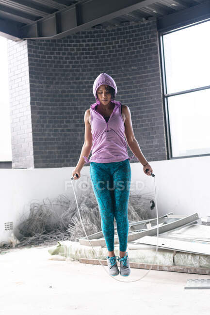 Femme afro-américaine portant un sweat à capuche sautant la corde dans un bâtiment urbain vide. forme physique urbaine mode de vie sain. — Photo de stock